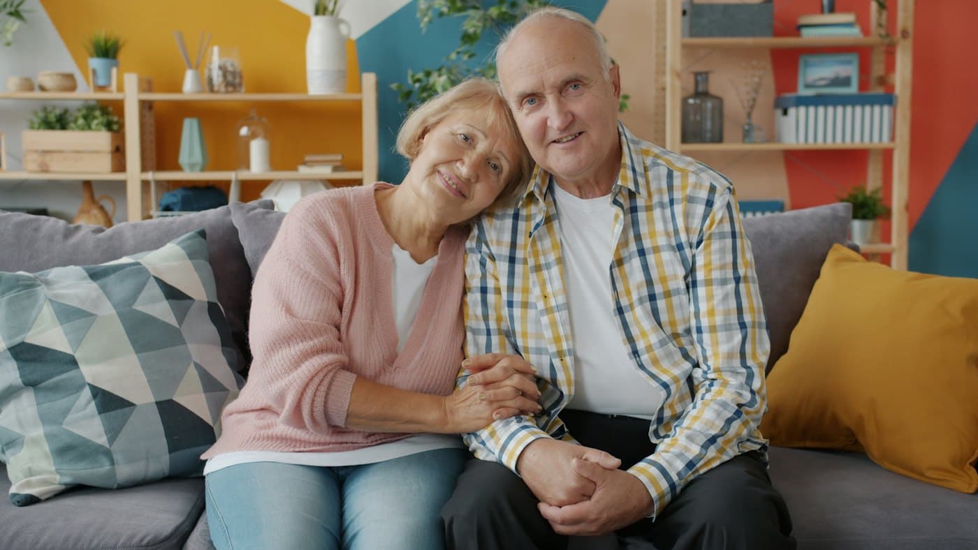 An older couple smiling and holding hands on their sofa