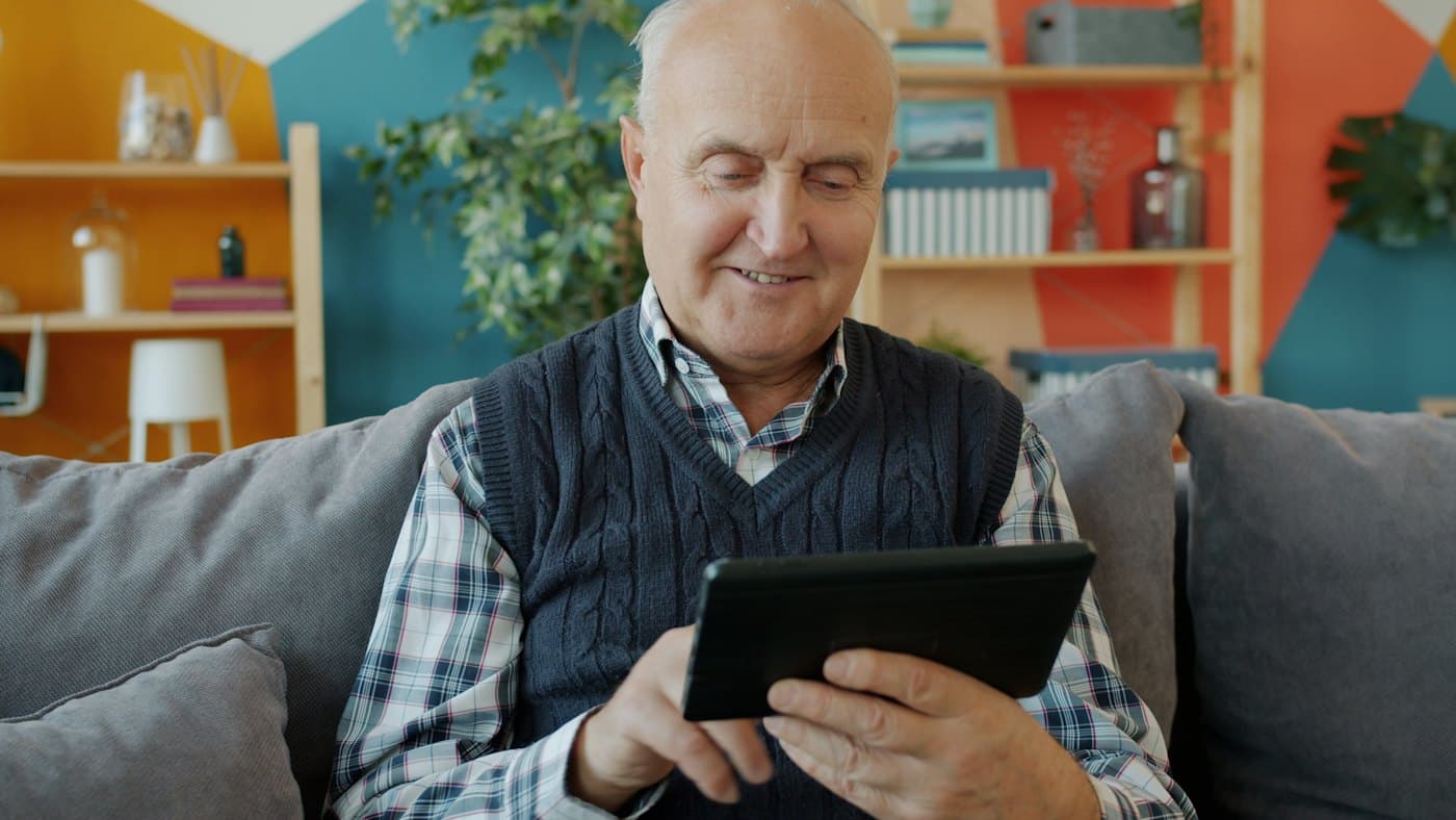 An older man smiling while using a tablet on his sofa